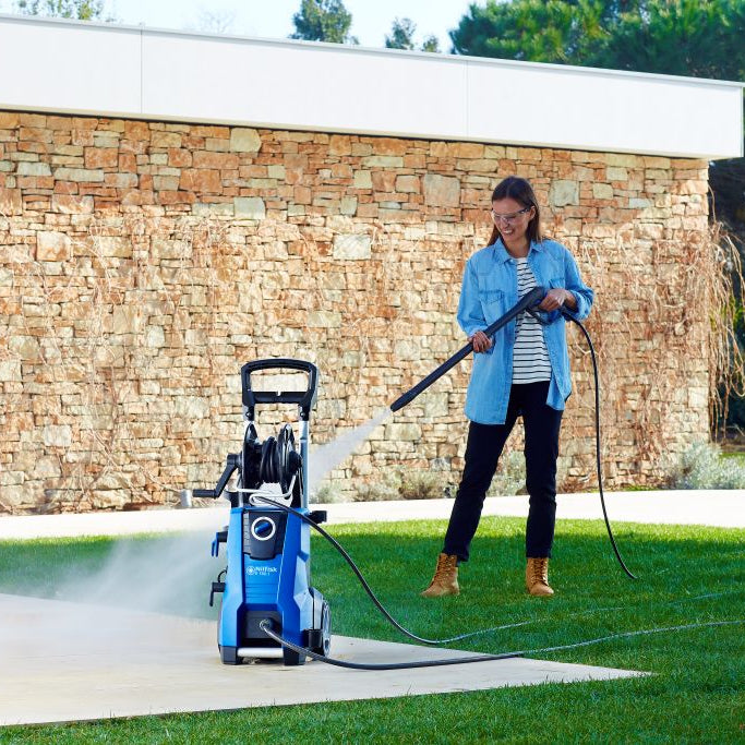 Blue pressure washer by Nilfisk in a yard, with woman in denim shirt operating it.
