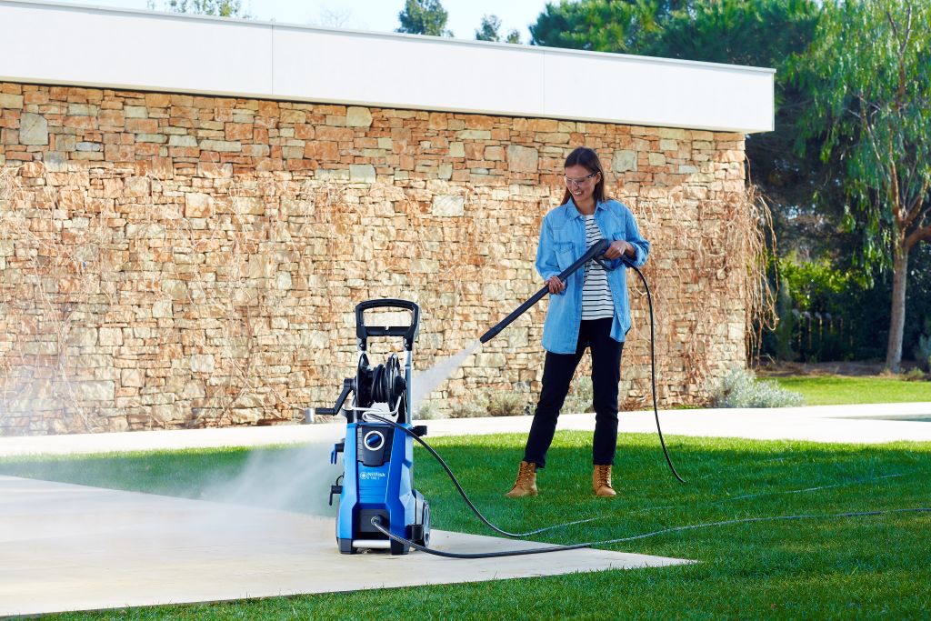 Blue pressure washer by Nilfisk in a yard, with woman in denim shirt operating it.
