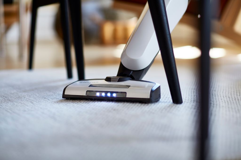 White cordless stick vacuum cleaner by Nilfisk, seen cleaning a light beige rug under black table legs.
