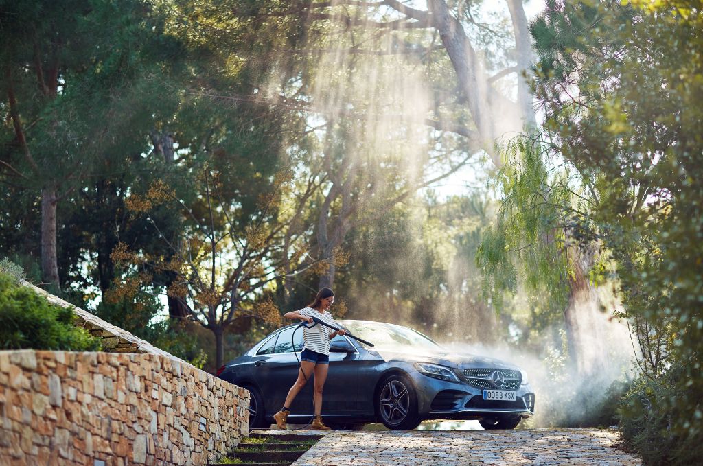 High-pressure washer for car washing by Nilfisk, with water spraying on a car, amidst green trees.