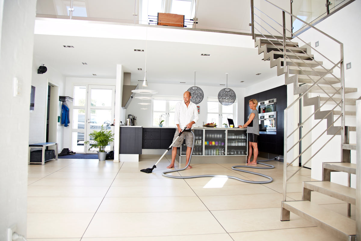 Central vacuum cleaner, wireless, by Performer, being used in a modern kitchen with sleek cabinets and steel staircase.