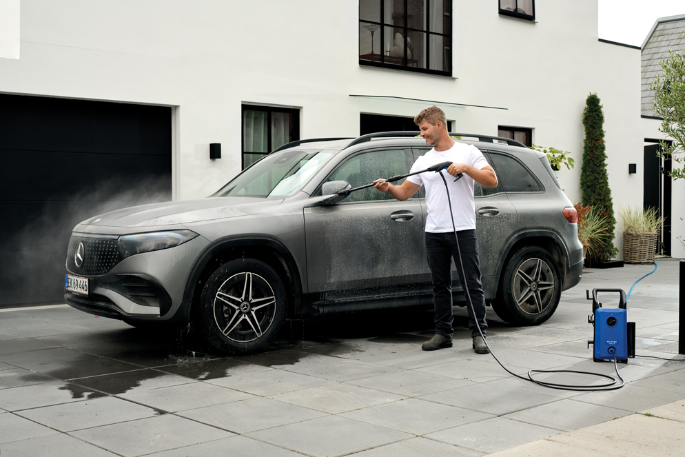 Blue compact high-pressure washer by Nilfisk in outdoor driveway use, spraying water on gray SUV, with black hose and wand, lifestyle side view