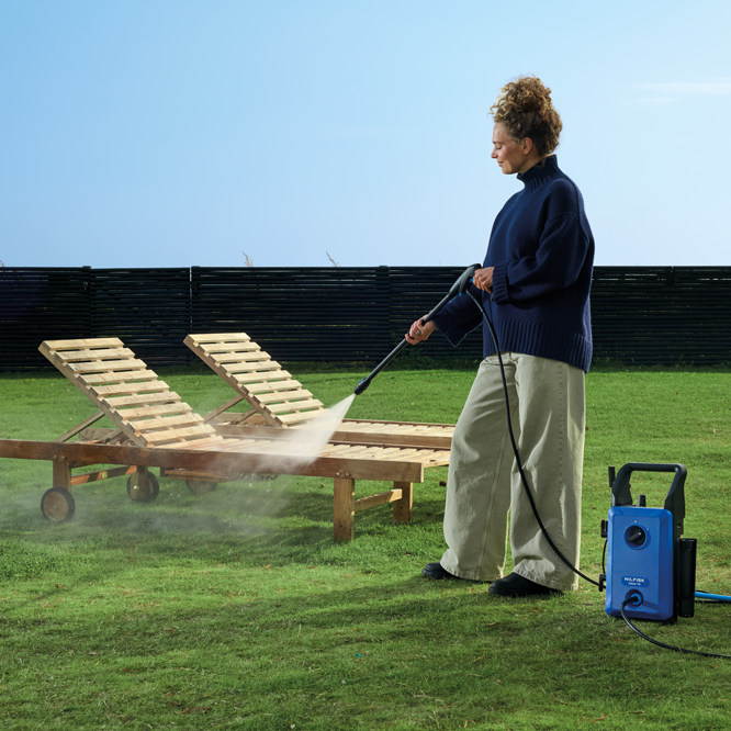 Blue high-pressure washer with black handle and hose, used outdoors on grass to clean wooden lounge chairs, side view lifestyle shot