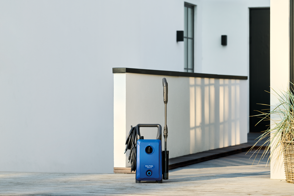 Blue high-pressure washer with black hose and spray lance by Nilfisk, positioned on wooden deck outdoors, side view.