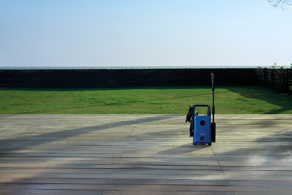 Blue high-pressure washer with black handle and hose, positioned on wooden deck, outdoor lifestyle view