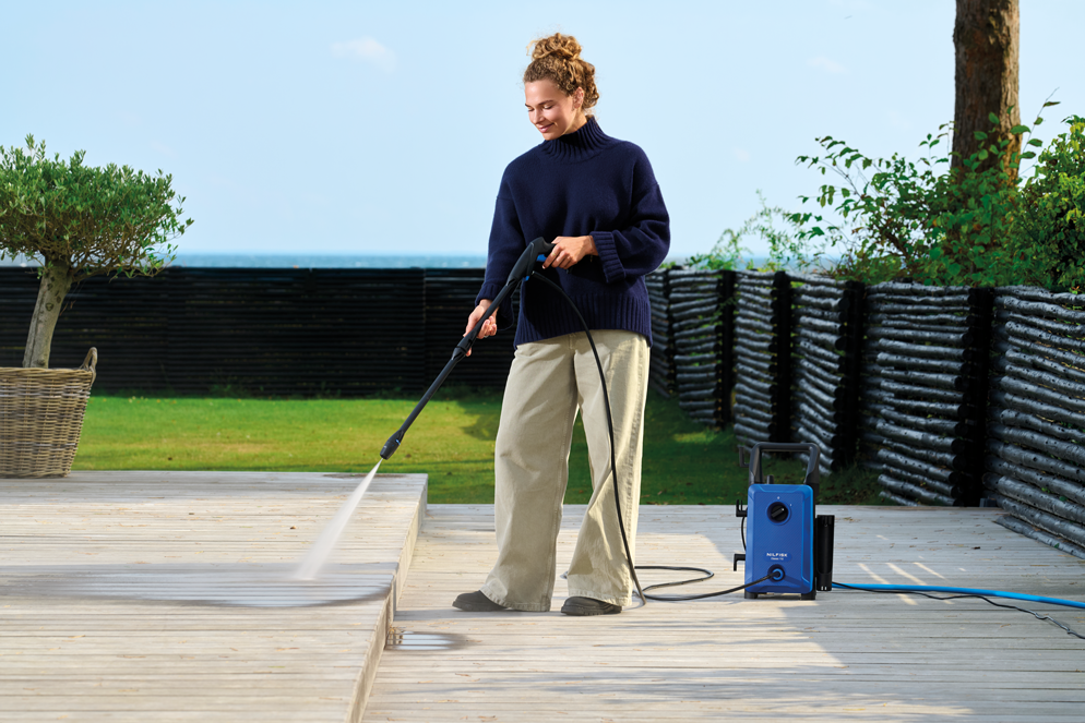 Blue high-pressure washer with black hose and wand, by Nilfisk, in outdoor garden setting, cleaning wooden deck, side view.