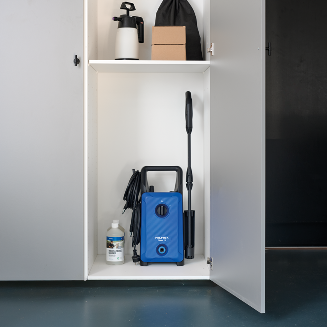 Blue high-pressure washer with black handle and hose, by Nilfisk, stored in white cabinet with cleaning accessories, front view.