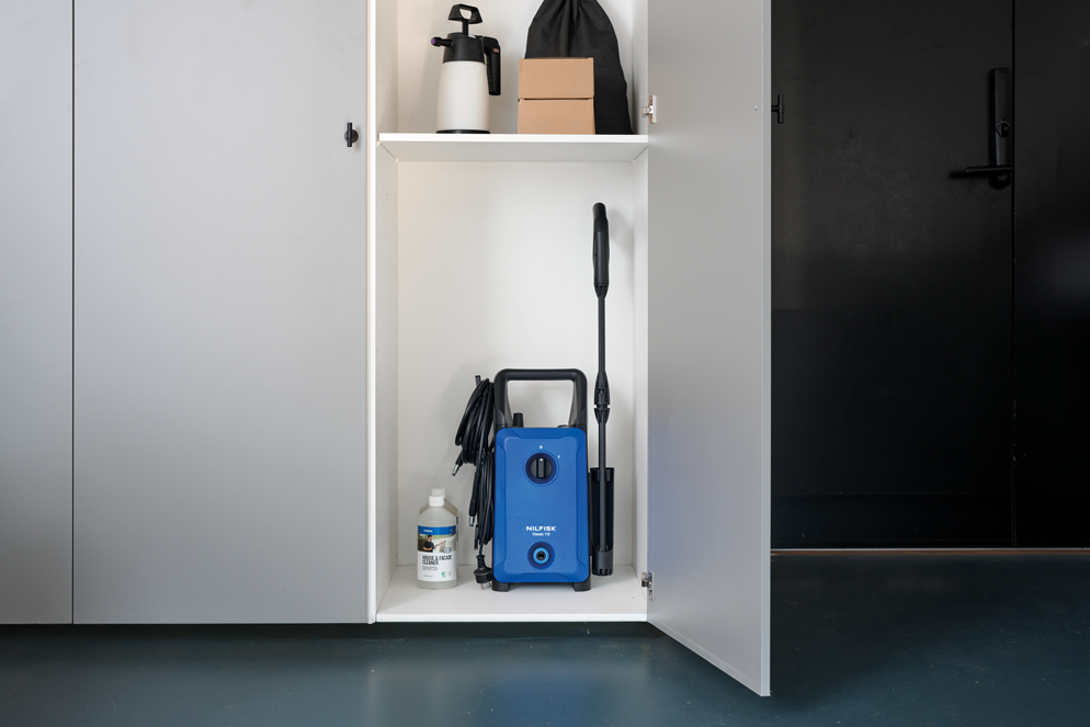 Blue high-pressure washer with black handle and hose, by Nilfisk, stored in white cabinet with cleaning accessories, front view.