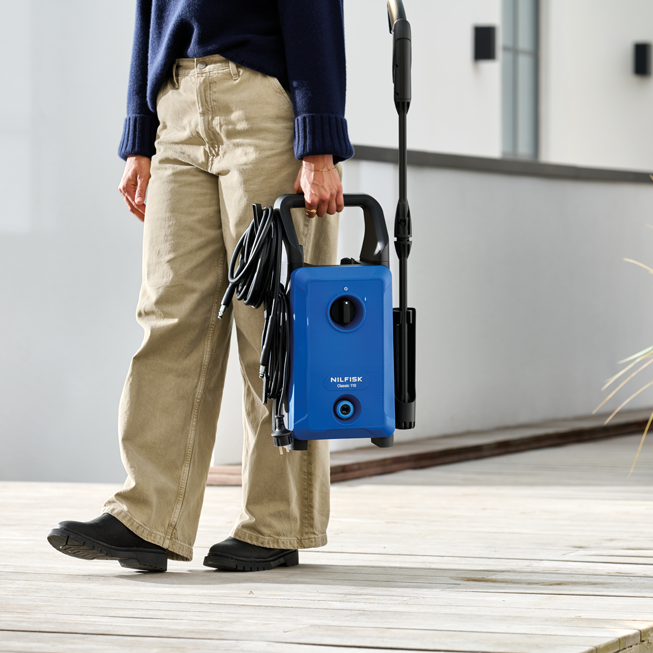 Blue portable high-pressure washer by Nilfisk, held by person in beige pants and navy sweater on wooden deck, side view.