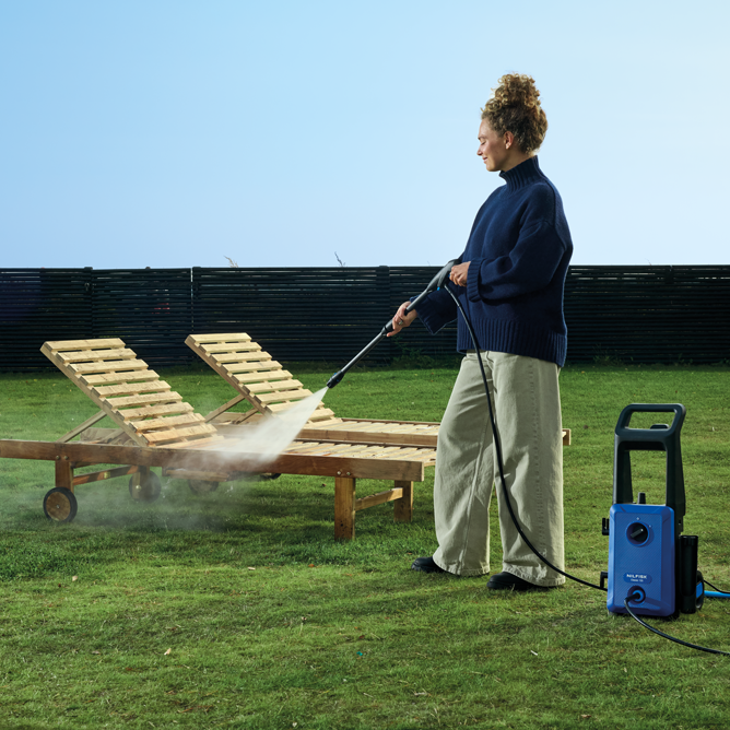 Blue and black high-pressure washer by Nilfisk in outdoor garden setting, used to clean wooden lounge chairs, side view with spray mist visible