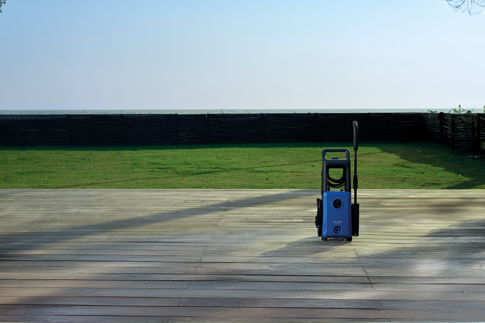 Blue and black high-pressure washer on wooden deck, side view, with coiled hose and spray gun, outdoors near grass and fence.