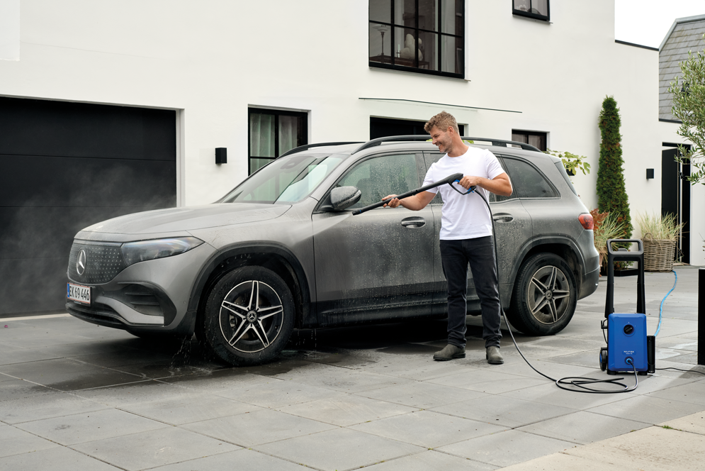 Blue portable high-pressure washer by Classic 140-6 in use, spraying water on a gray SUV in a driveway, side view with hose and wand.