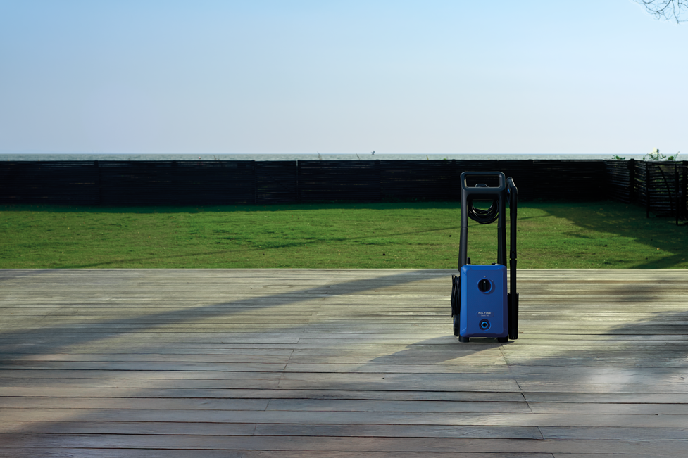 Blue electric pressure washer with black handle and hose, by Nilfisk, on wooden deck outdoors, side view, sunny day