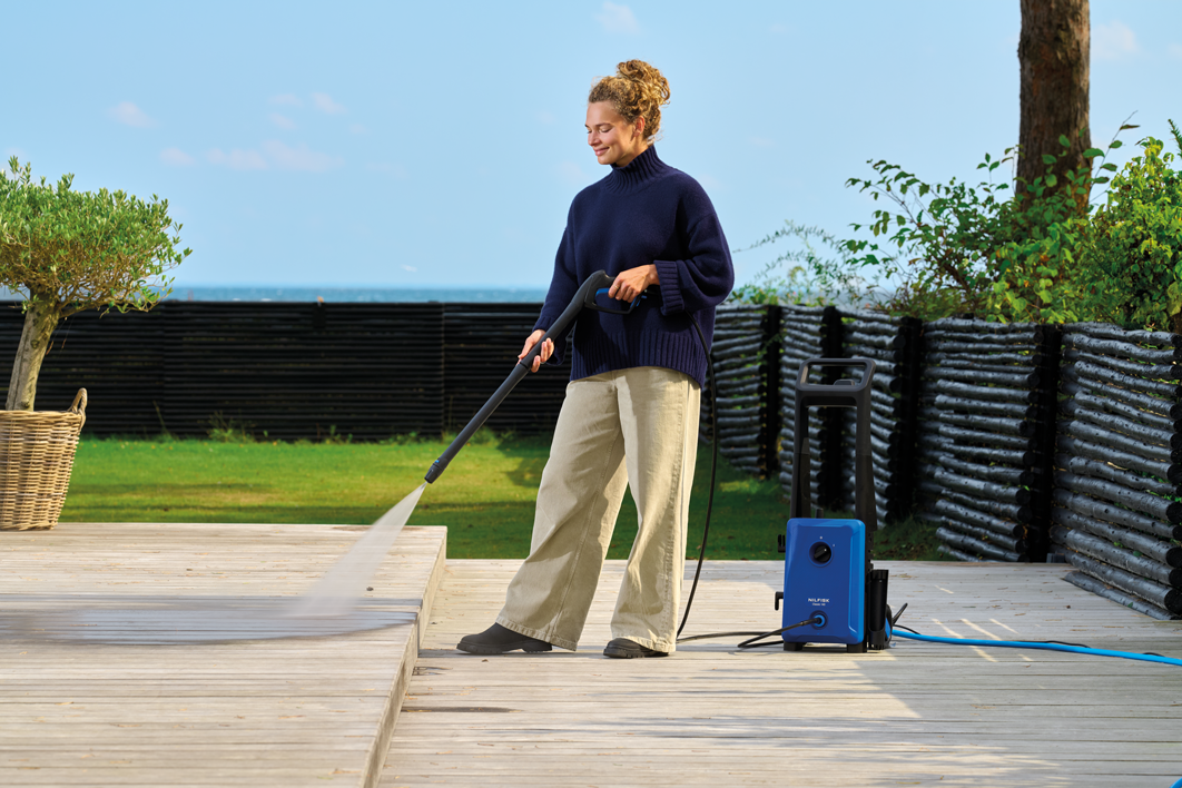 Blue high-pressure washer with black handle and hose, by Nilfisk, in outdoor lifestyle use on wooden deck, side view