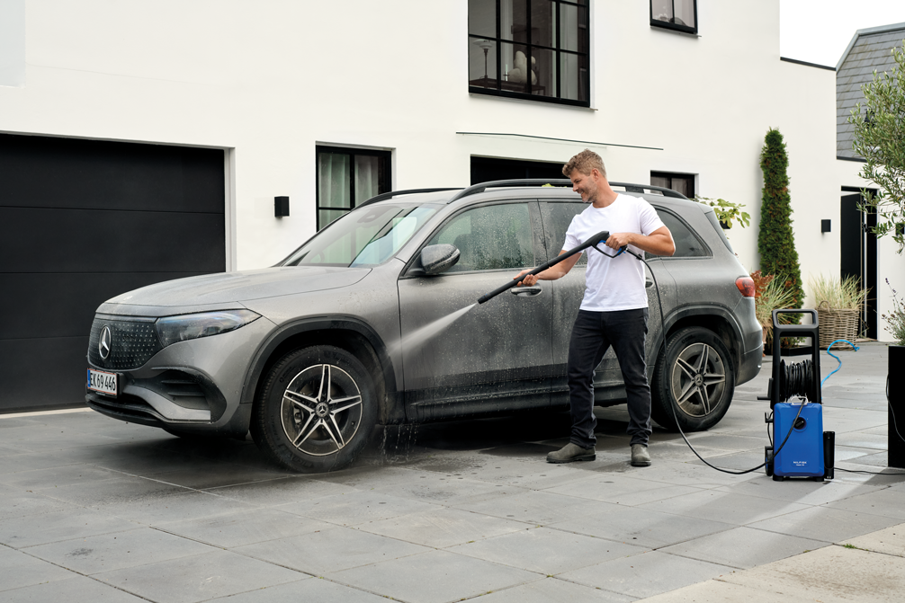 Blue portable high-pressure washer by Classic 150-8 in use, spraying water on a gray SUV in a driveway, side view with coiled hose and handle.