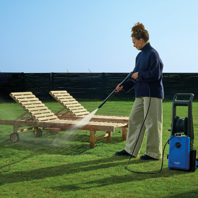 Blue high-pressure washer with black hose reel and handle, by Holmen, used outdoors on wooden lounge chairs in a grassy yard.