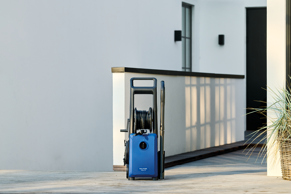 Blue high-pressure washer with black hose reel and spray lance by Nilfisk, positioned on outdoor patio, side view, modern design.