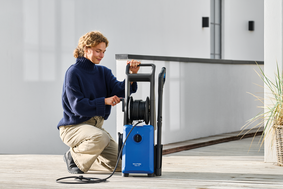 Blue and black high-pressure washer with hose reel by Nilfisk, shown on wooden deck with person in navy sweater and beige pants kneeling beside it.