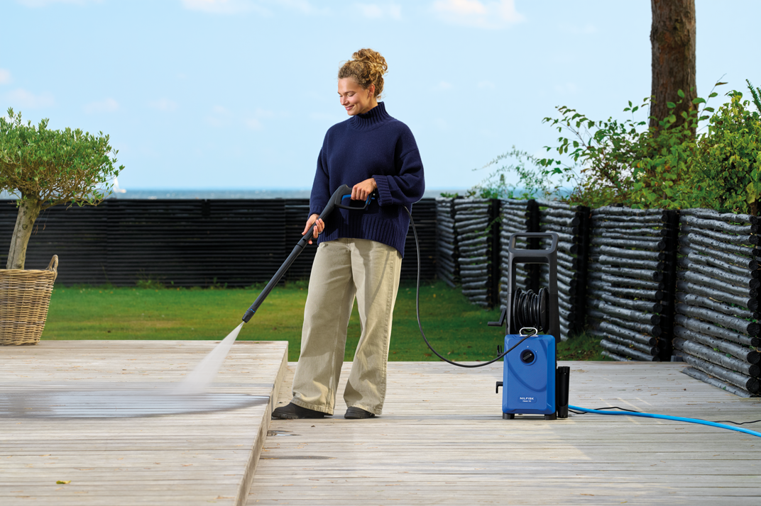 Blue high-pressure washer with black hose reel and handle, by Nilfisk, on wooden deck with woman using spray wand, outdoor lifestyle view