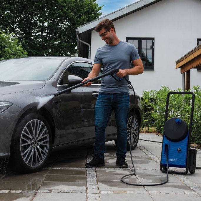 Blue and black high-pressure car washer by Core 150 in use, spraying a gray sedan in a driveway, side view.