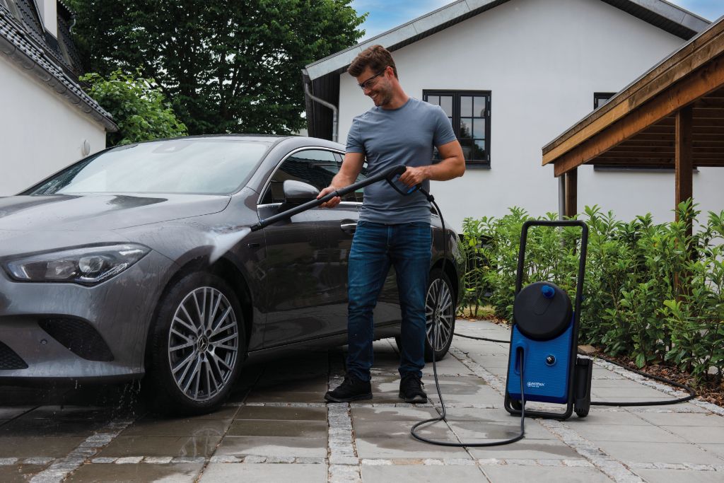 Blue and black pressure washer by Nilfisk, with a man washing a car outside a house.
