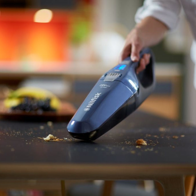 Blue stick vacuum cleaner by Nilfisk cleaning breadcrumbs on a dark table, with fruit in the background.