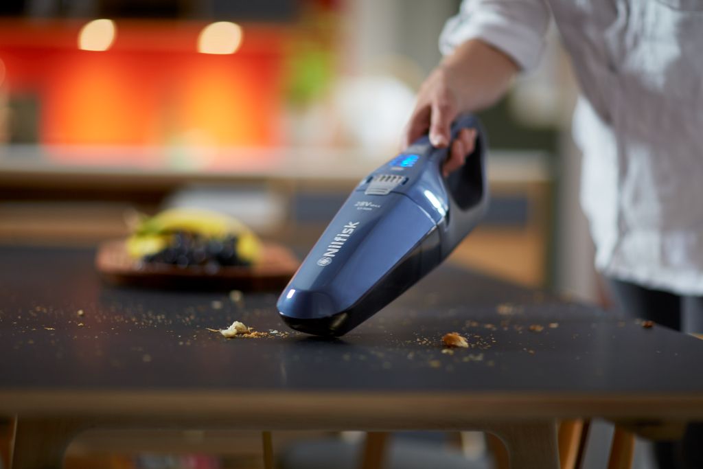 Blue stick vacuum cleaner by Nilfisk cleaning breadcrumbs on a dark table, with fruit in the background.