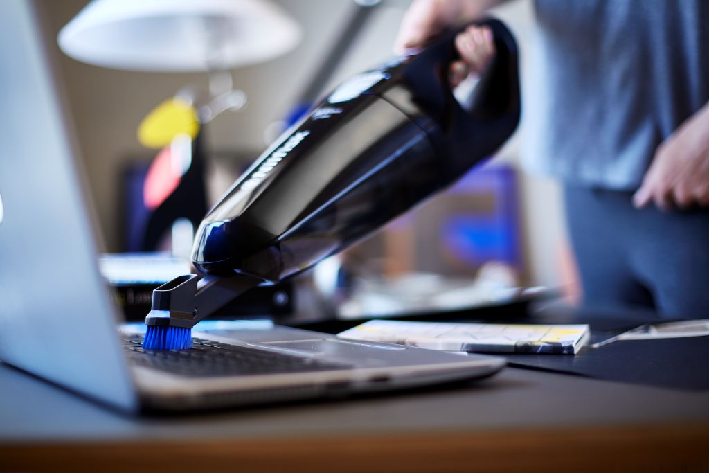 Black handheld vacuum cleaner, by VONROC, cleaning a silver laptop with a brush attachment.