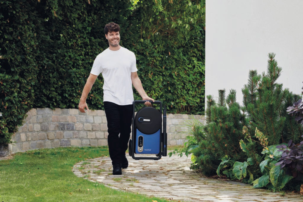 Blue pressure washer by Nilfisk, with hose reel and black frame, being carried on a brick pathway.